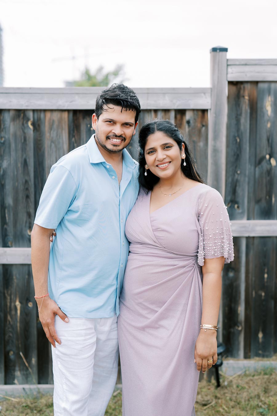 Couple smiling and posing together in front of a wooden fence; man in light blue shirt and white pants, woman in light purple dress.