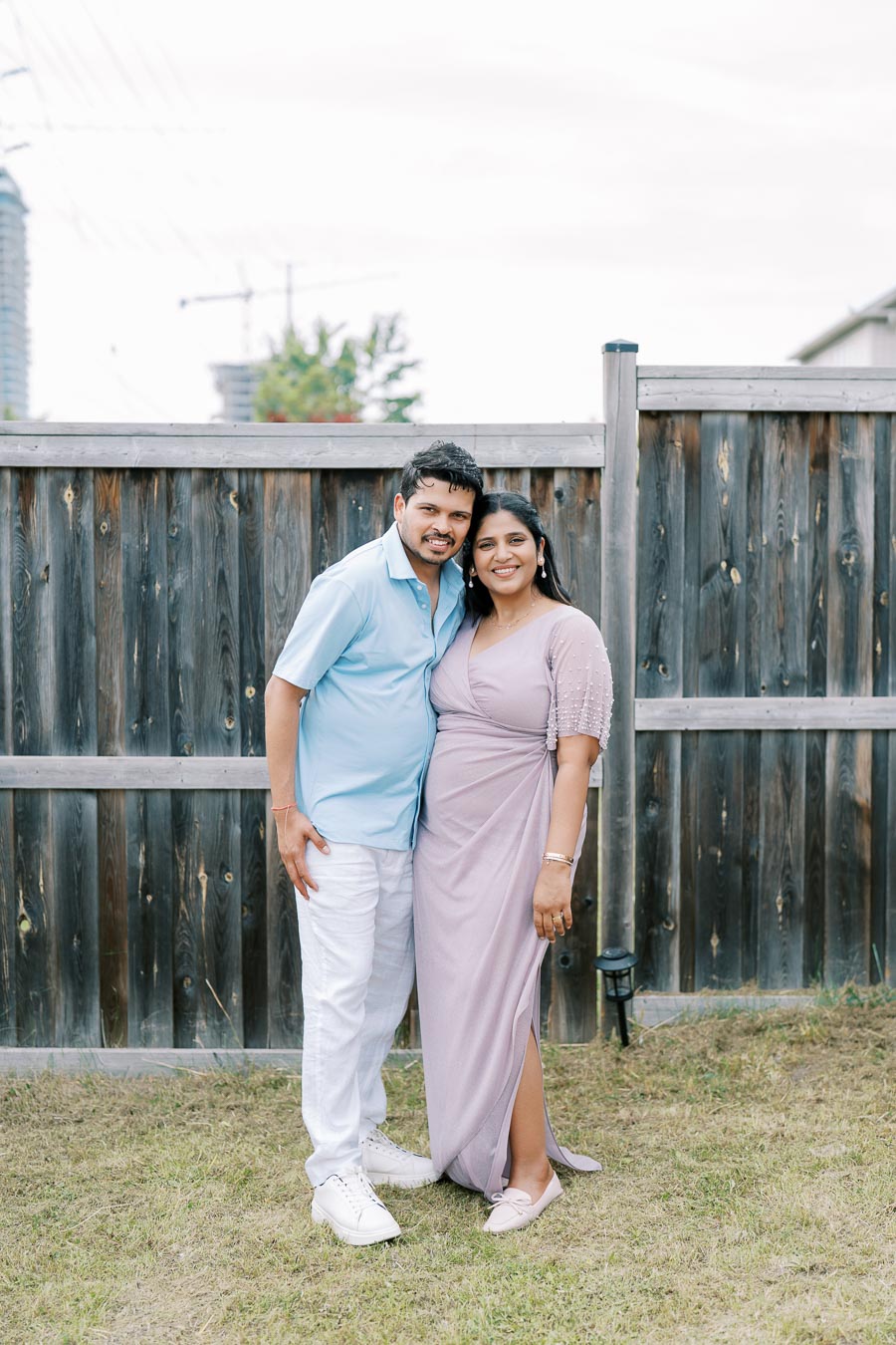 A smiling couple poses in front of a wooden fence in a backyard. The woman is wearing a lavender wrap dress, and the man is in a light blue shirt and white pants. The background features a partly cloudy sky and a hint of city buildings.