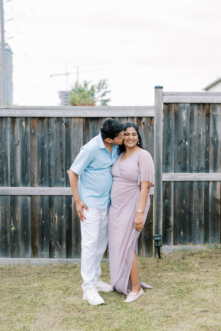 A couple shares a tender moment, standing in front of a wooden fence in a garden setting. The man, wearing a light blue shirt and white pants, kisses the smiling woman, who is dressed in a light purple dress.