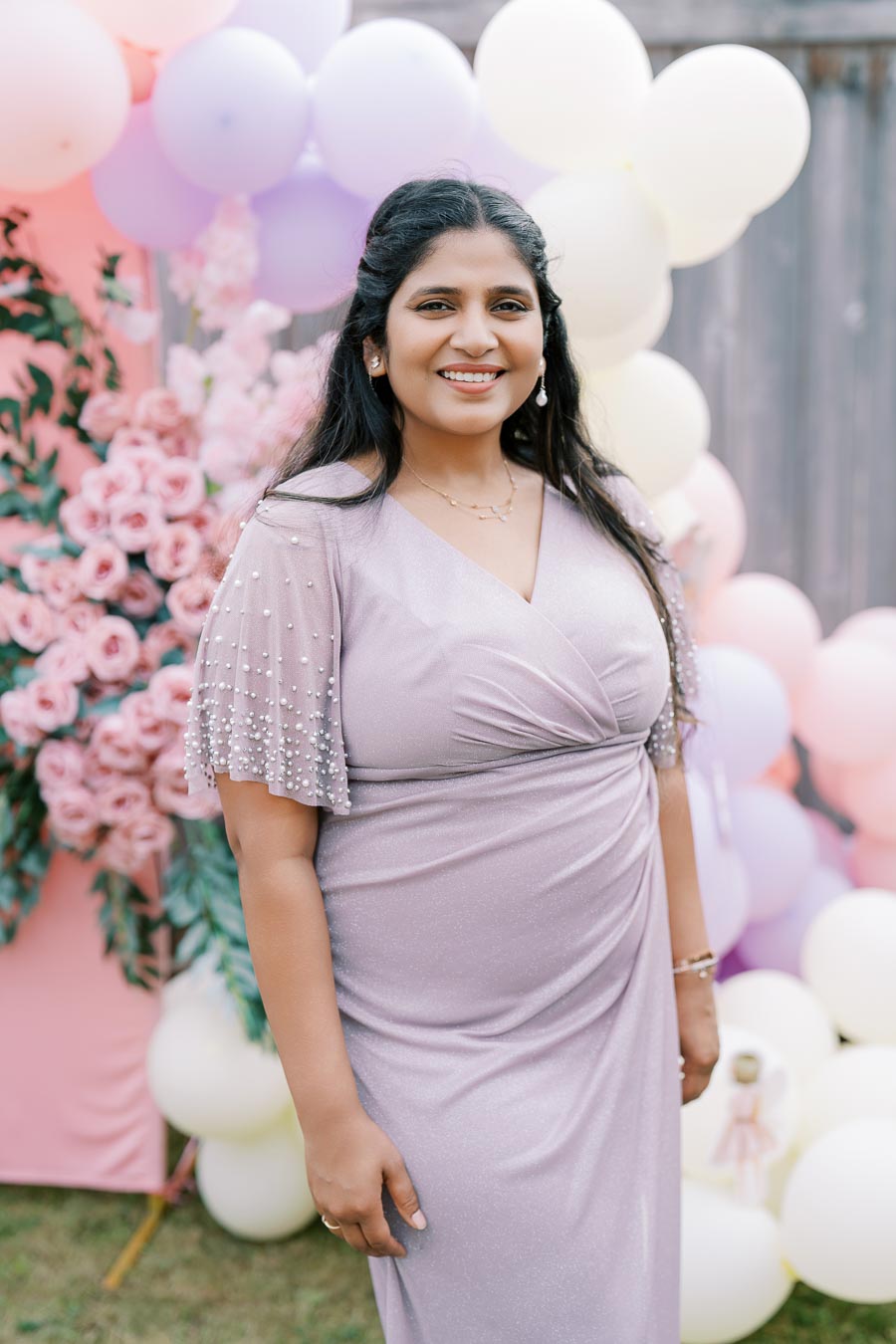 Smiling woman in a lavender dress at a garden party, surrounded by a colorful balloon and flower backdrop.