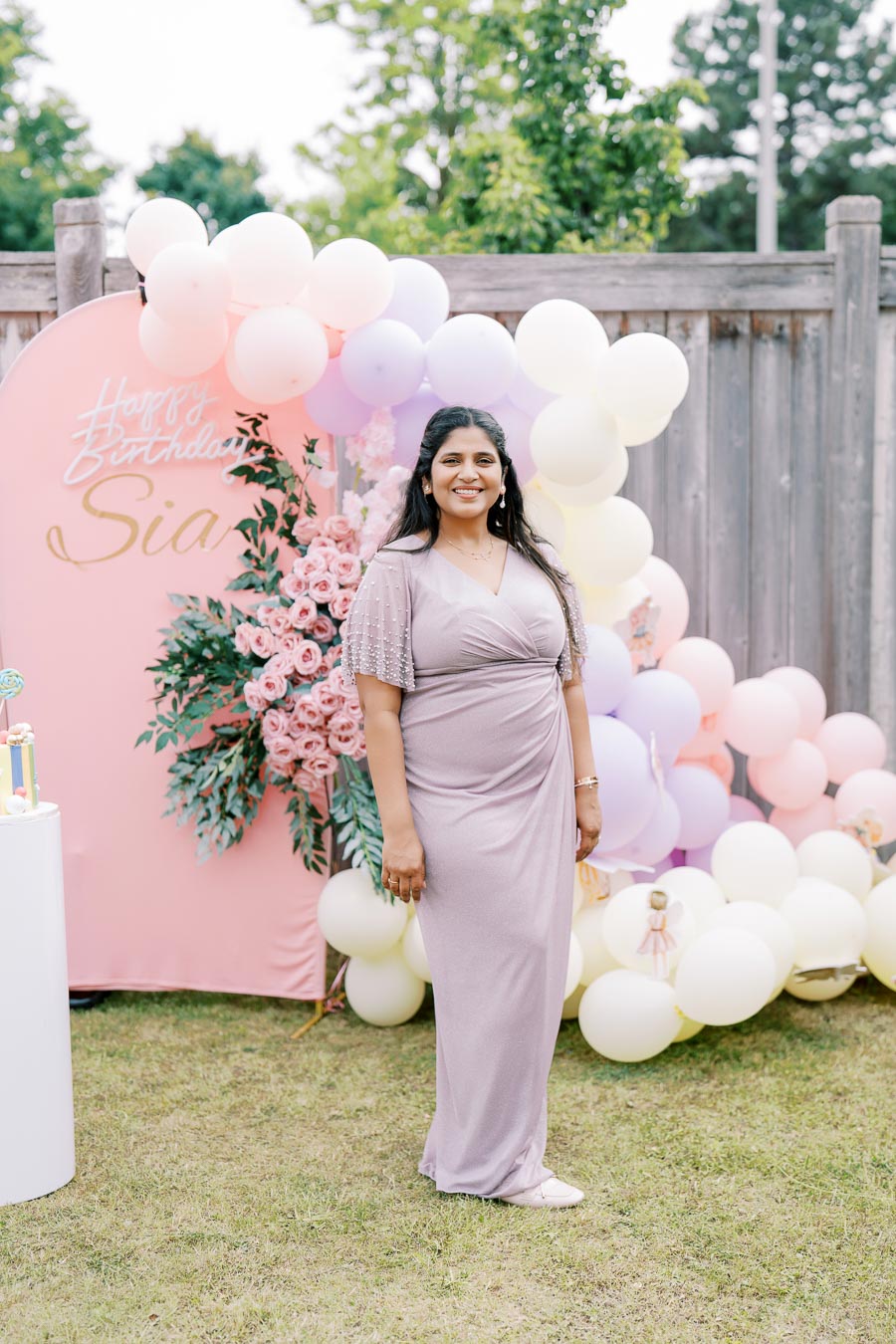 Woman in a lavender dress smiling at an outdoor birthday celebration, surrounded by pastel balloons, flowers, and a pink backdrop with Happy Birthday text.