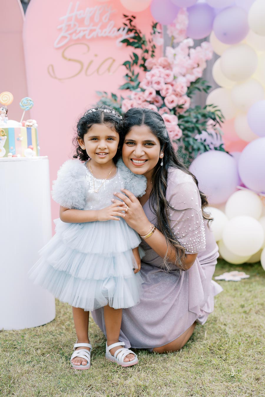 A woman and a young girl smiling at a birthday party, surrounded by colorful balloons and floral decorations. The girl is wearing a light blue ruffled dress, and the woman is in a sparkling outfit, both kneeling on grass near a festive cake.