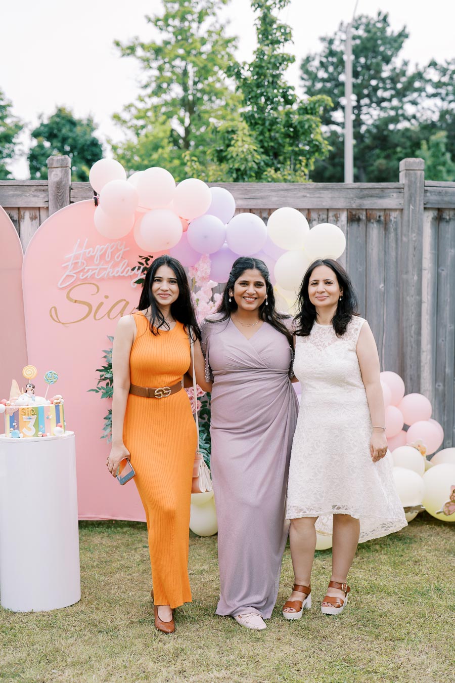 Three women smiling at a birthday party, standing in front of a colorful balloon backdrop and a Happy Birthday sign outdoors, with a decorated cake nearby.