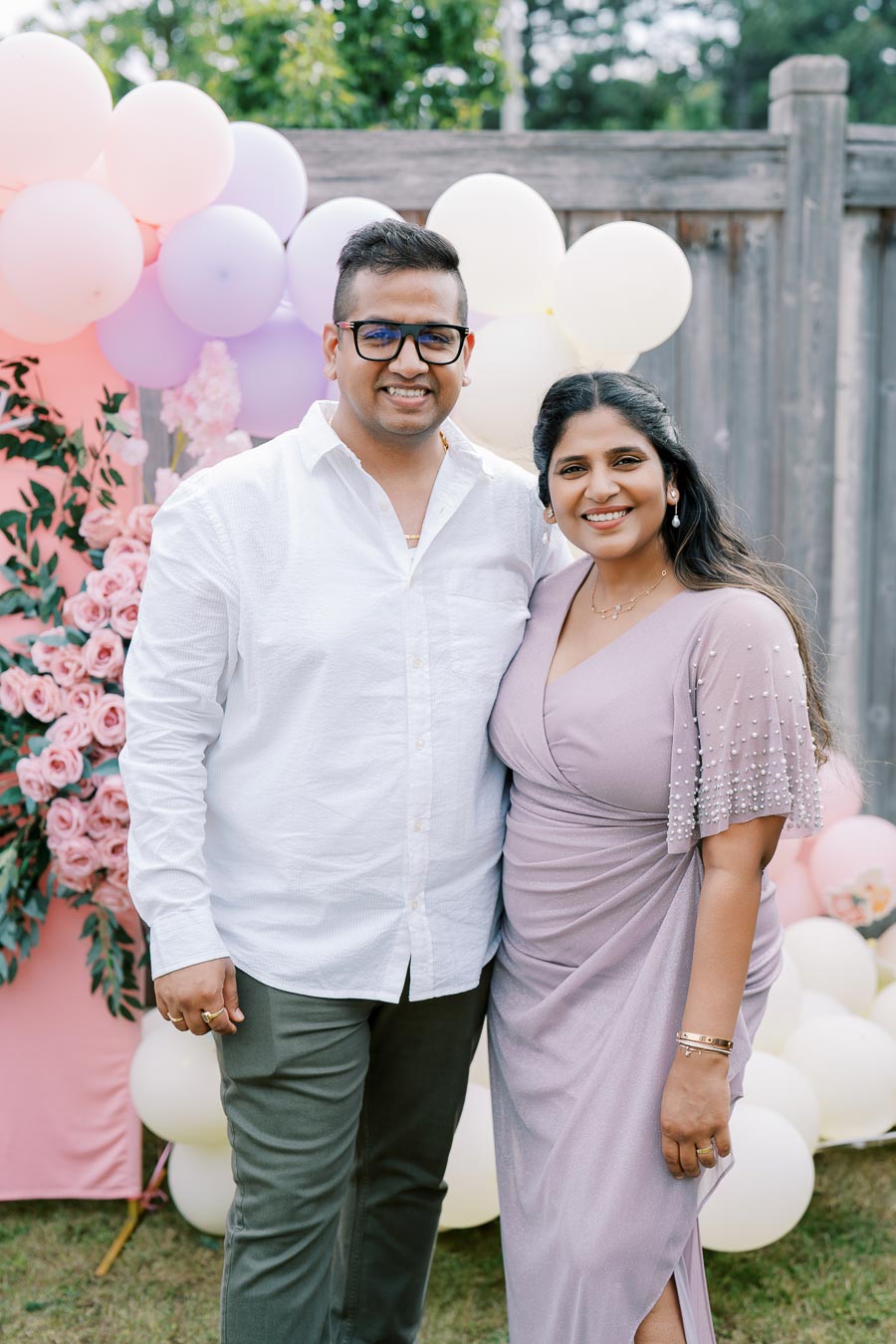 A couple smiling at an outdoor event, surrounded by pastel balloons and pink floral decorations, with a wooden fence and greenery in the background