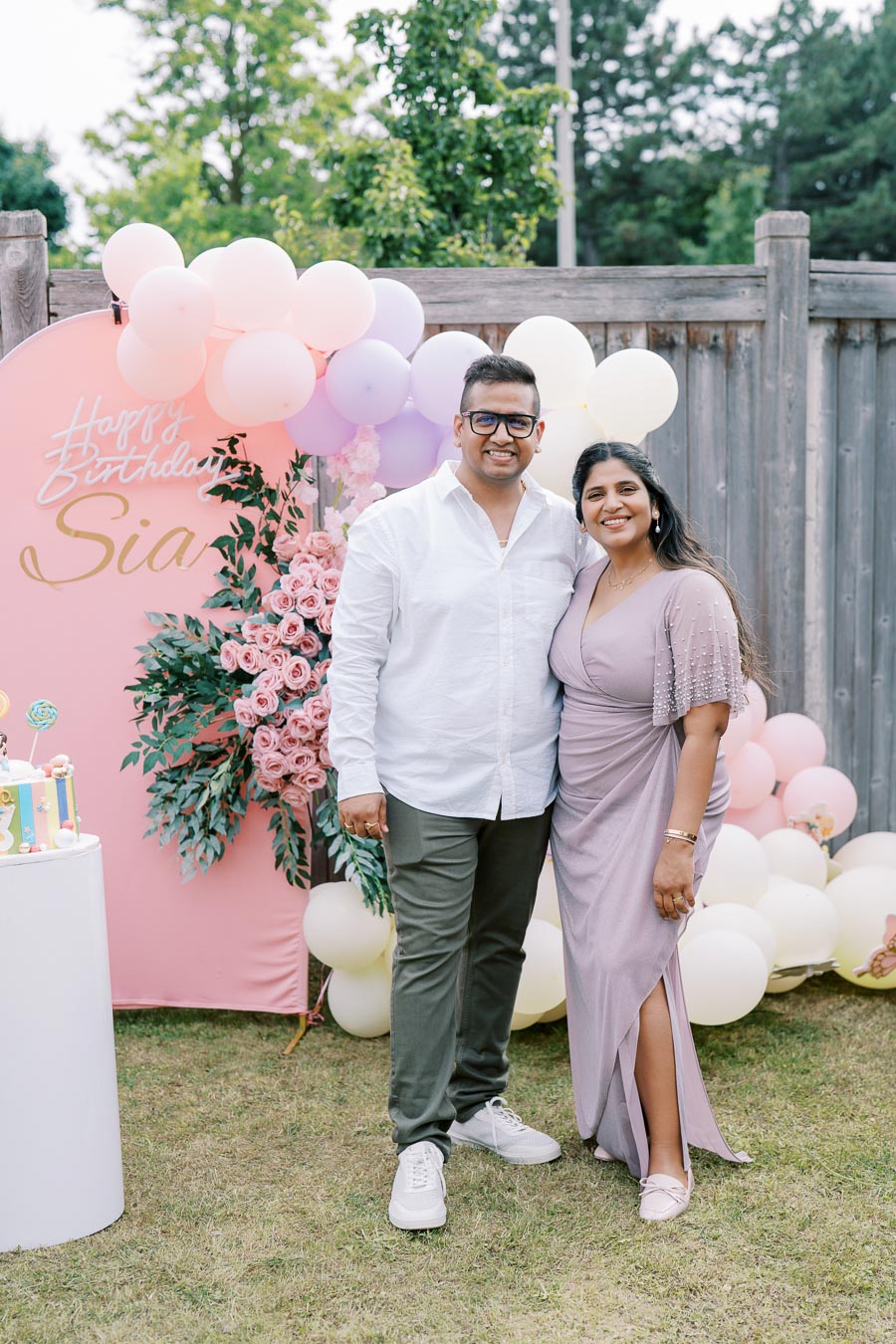 A smiling couple posing in front of a colorful birthday backdrop with balloons and flowers, celebrating a special occasion outdoors.