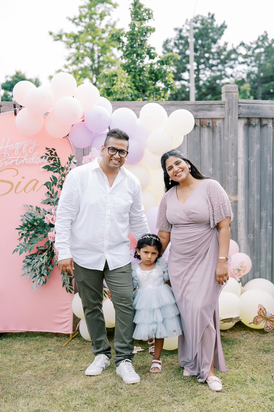 A family celebrating a child's birthday outdoors, featuring a woman in a light purple dress, a man in a white shirt with green pants, and a young girl in a blue dress. They stand in front of a decorative setup with balloons and a birthday banner on a pink backdrop.