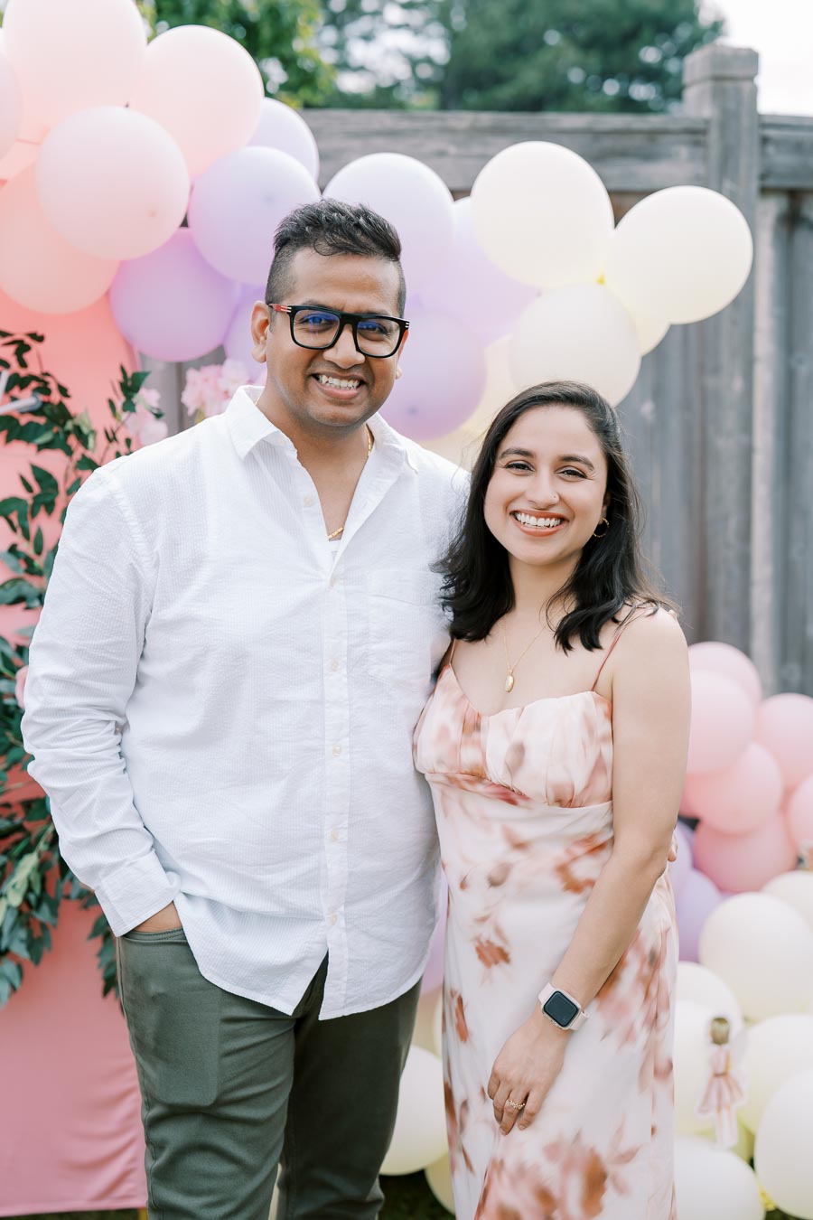 A smiling couple posing in front of a colorful balloon backdrop at an outdoor celebration. The man is wearing a white shirt and glasses, and the woman is in a floral dress with a smartwatch. Lush greenery is visible in the background.