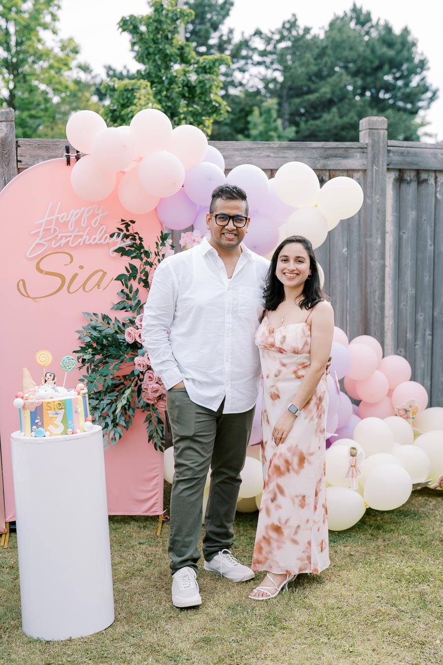 Couple posing at a child's third birthday party, featuring a colorful cake, festive balloons, and a Happy Birthday Sia sign in an outdoor setting.