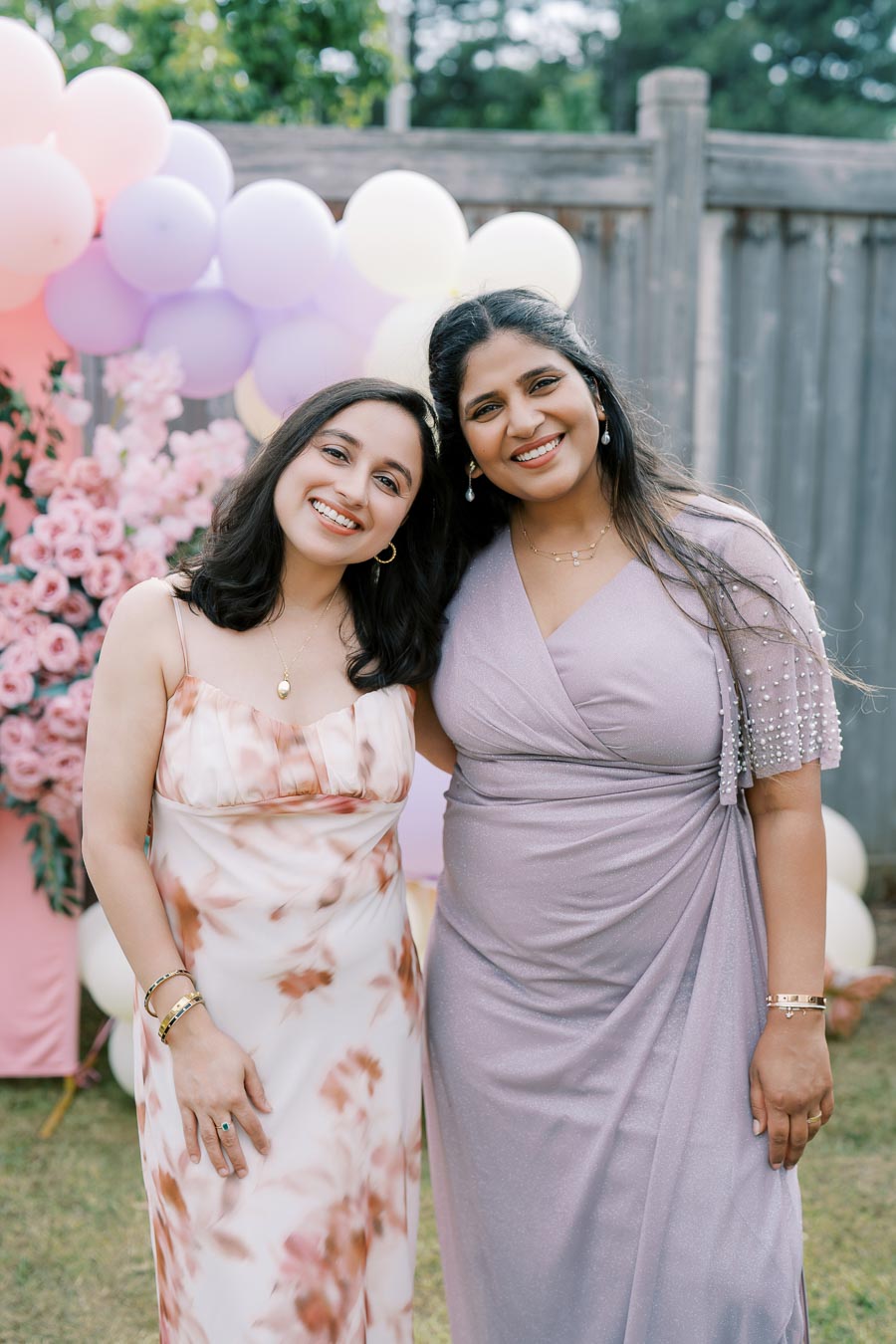 Two women smiling at an outdoor event with a backdrop of pastel balloons and pink flowers