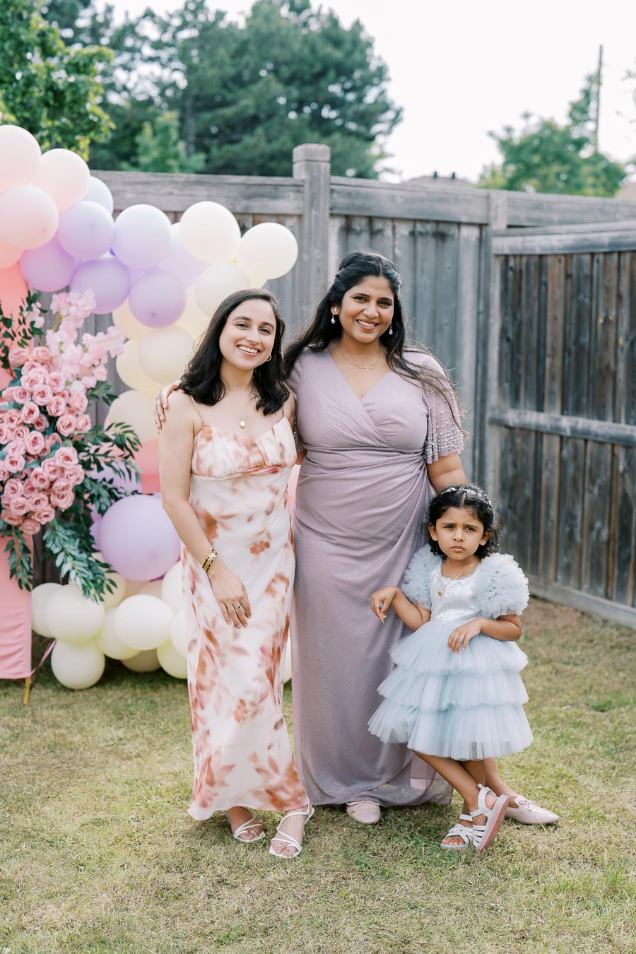 A joyful outdoor celebration with two women and a young girl dressed in elegant attire, standing in front of a colorful balloon decoration and flowers, set against a wooden fence and greenery.