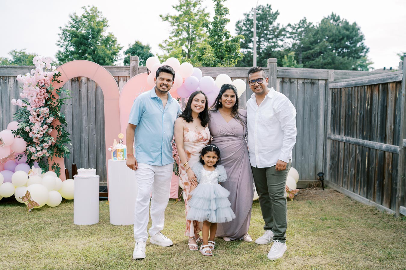 A family gathering celebrating a child's birthday party in a backyard setting, featuring five people smiling in front of a decorative backdrop with balloons and flowers.
