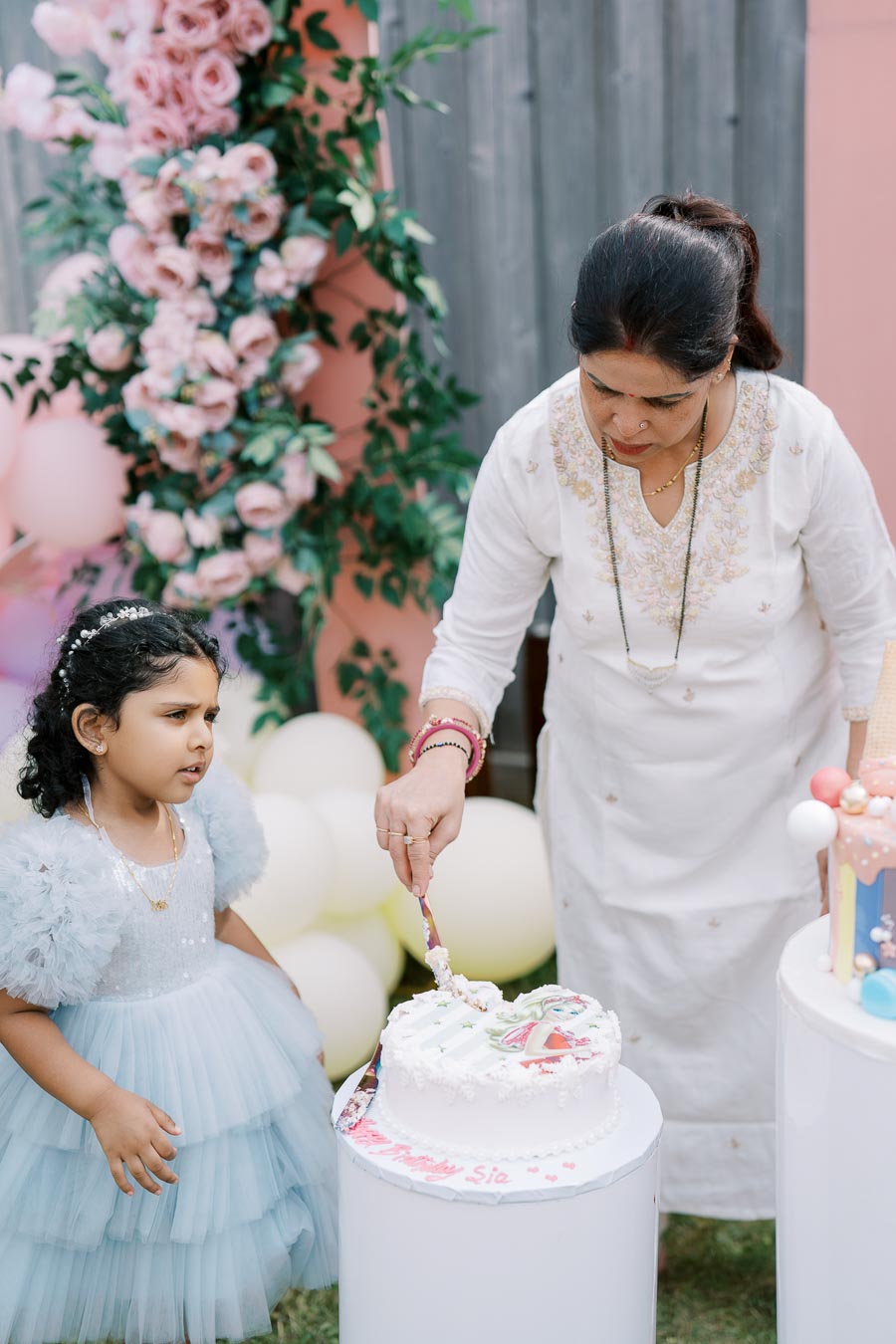 A child in a light blue dress watches an adult in a white embroidered outfit cut a birthday cake at an outdoor celebration, surrounded by pink floral decorations and balloons.