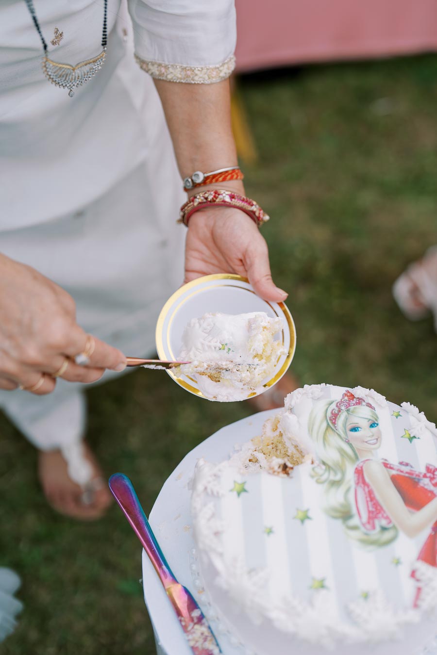 A woman in traditional attire serving a slice of decorated birthday cake with a character image, holding a plate and using a colorful knife, captured at an outdoor celebration.