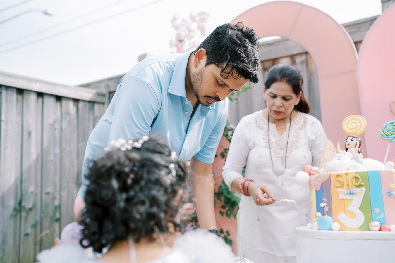 A man and a woman standing near a vibrant birthday cake with a number 3, at an outdoor celebration.