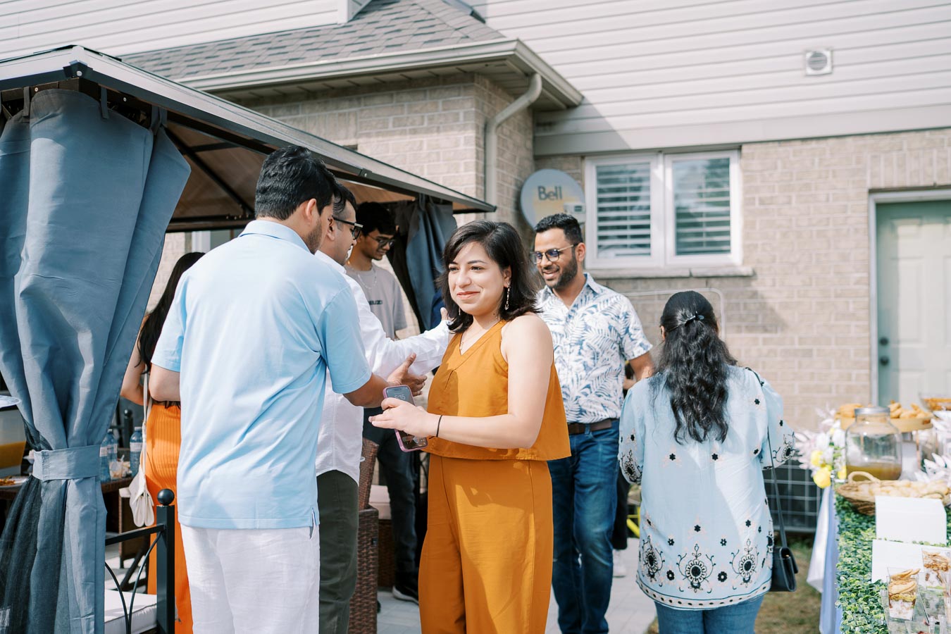 A group of people socializing at an outdoor gathering, with some chatting and shaking hands, set against the backdrop of a house with a shaded patio and a food table nearby.