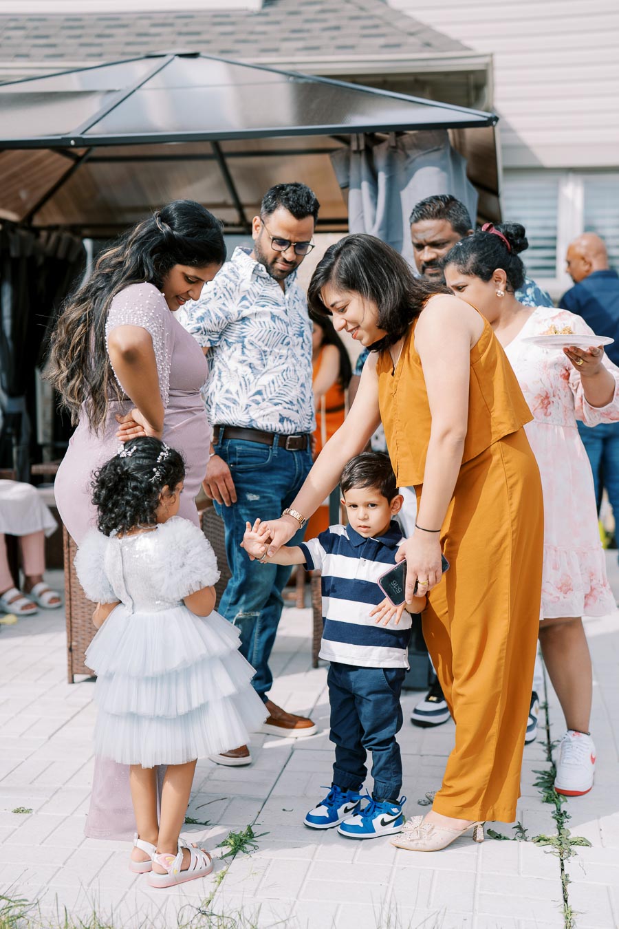 A group of people, including children and adults, gathered outdoors at a casual event or celebration. Two children are interacting with each other, while adults stand around them in conversation. The setting seems to be a backyard or patio with a gazebo in the background.