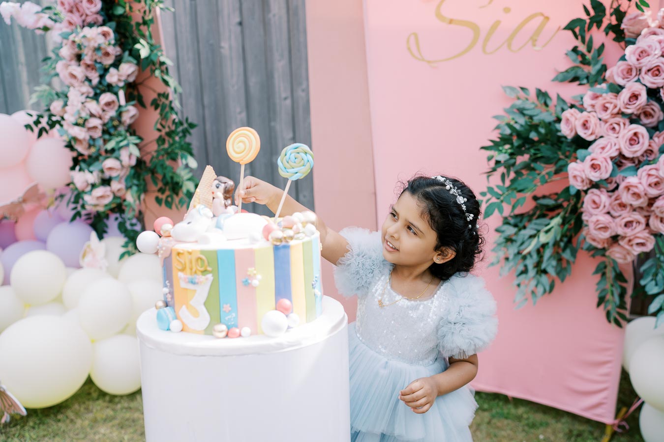 A young girl in a blue dress celebrates a birthday, reaching towards a colorful cake adorned with decorations and the number three, set against a festive backdrop of pink roses and balloons.