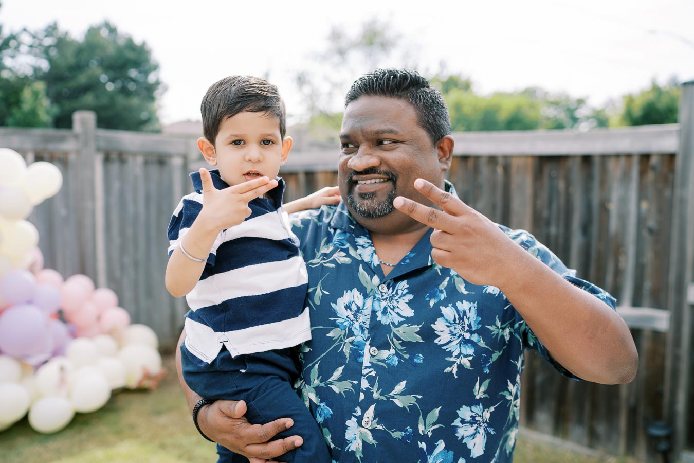 Father and son making peace signs in a backyard celebration with a balloon arrangement.