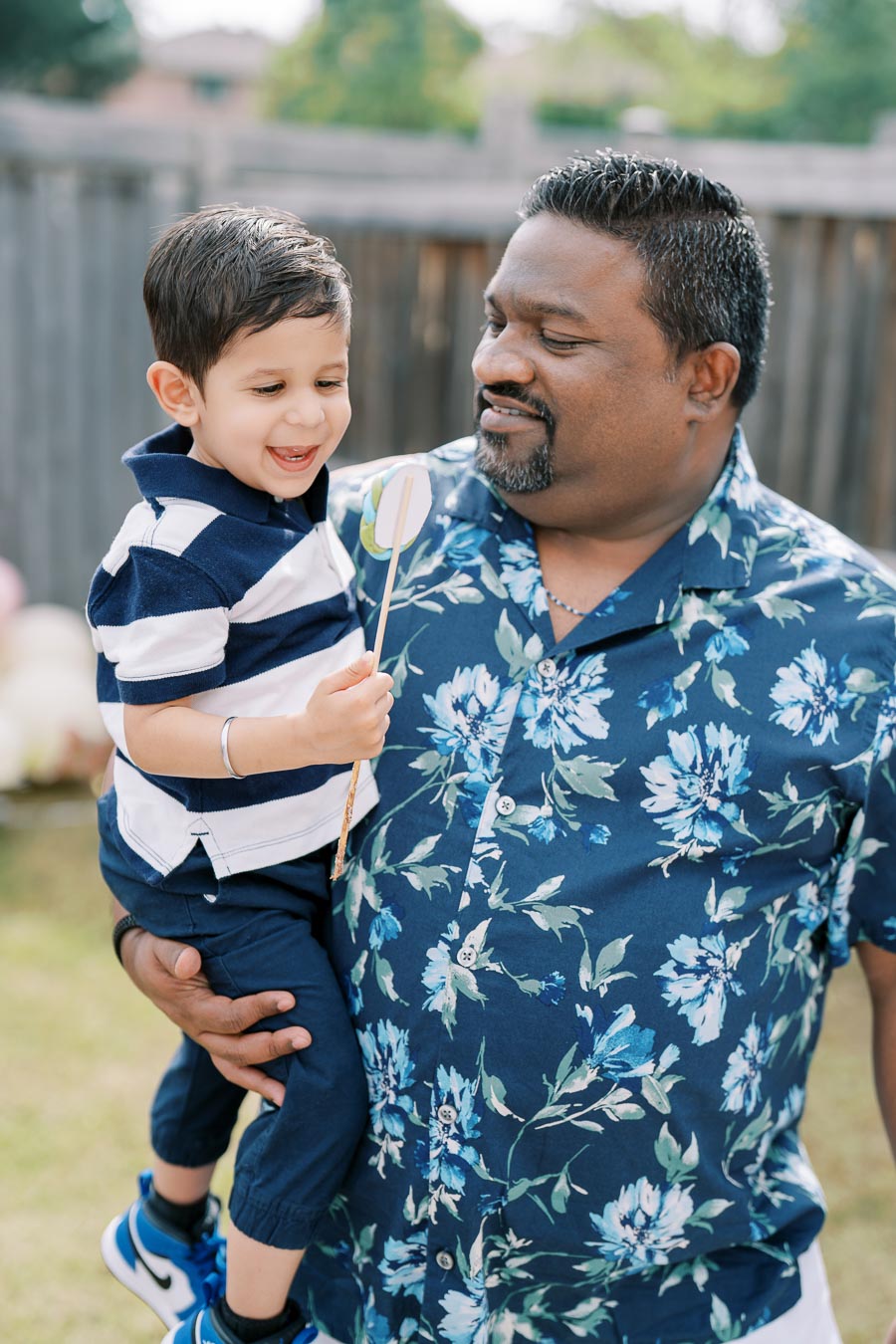 Smiling man holding toddler in backyard, child holding stick, both in floral and striped outfits, wooden fence and greenery in background, joyful outdoor moment.