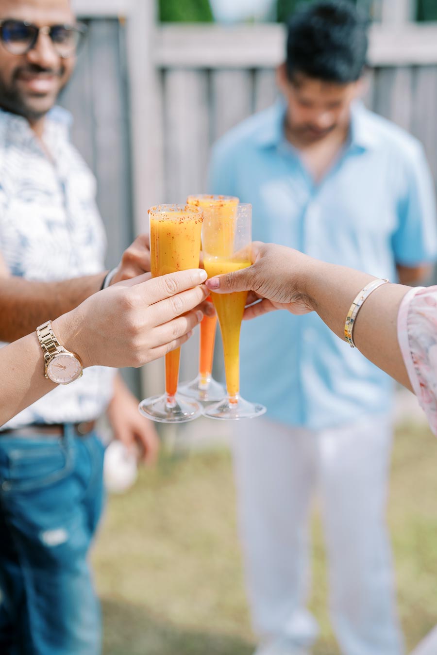 A group of people holding and toasting with colorful cocktails in clear glasses, outdoors. The drinks have a spicy rim, and the background features a blurred fence and person in casual attire, creating a festive and lively atmosphere.