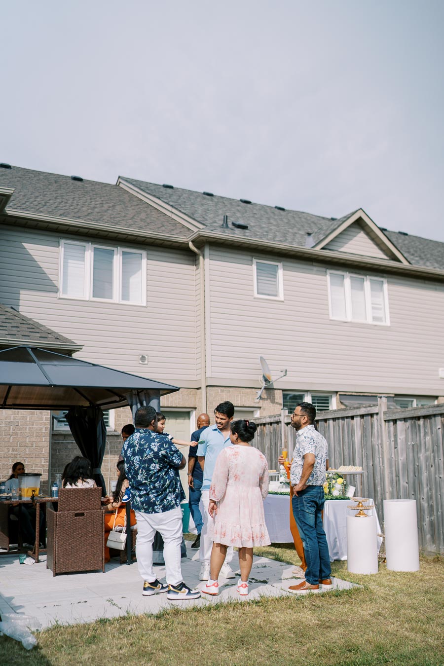 A group of people gathered in a backyard patio during a sunny outdoor celebration, with a table set up for refreshments and a modern townhouse in the background.