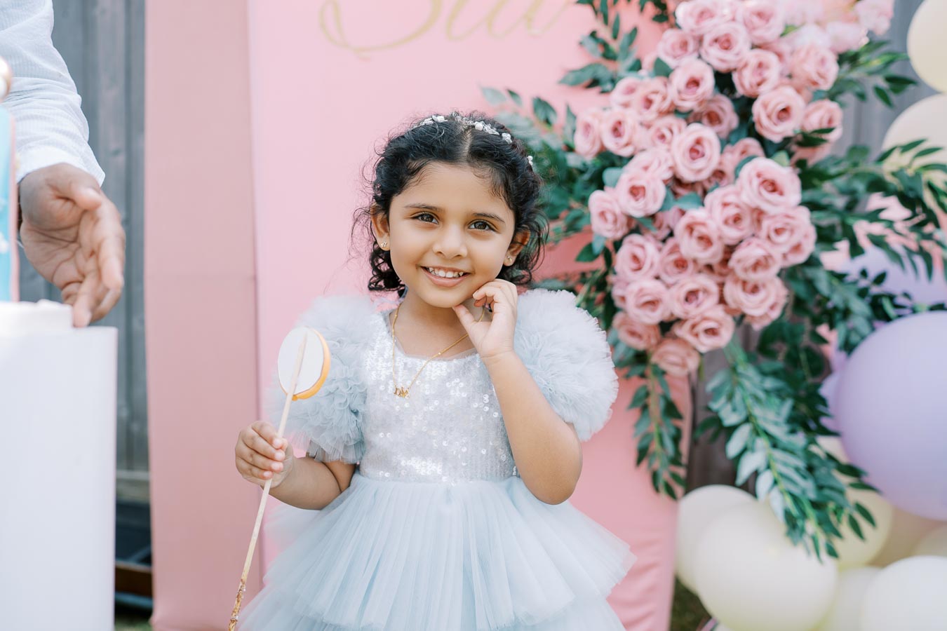 A young girl dressed in a blue, fluffy, sequined dress smiling at a birthday party, holding a small wand. She is standing in front of a pink backdrop adorned with lush pink rose decorations and pastel-colored balloons.