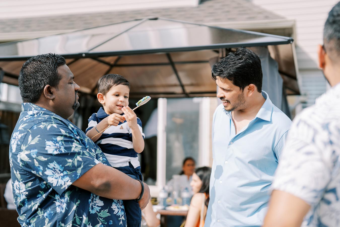 A man in a floral shirt holding a young boy who is showing a lollipop to another man in a light blue shirt, while standing outdoors near a gazebo during a sunny day.