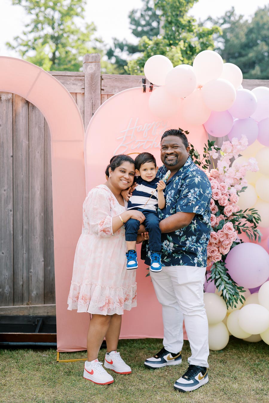 A smiling family celebrates a birthday outdoors, posing in front of a pink backdrop with balloons and flowers. The parents hold their young child, creating a joyful and festive atmosphere.
