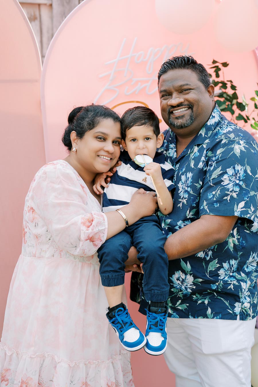 A smiling family of three celebrating a birthday. The parents are holding their young child in front of a pink backdrop with Happy Birthday written on it. The child is wearing blue sneakers and holding a lollipop.