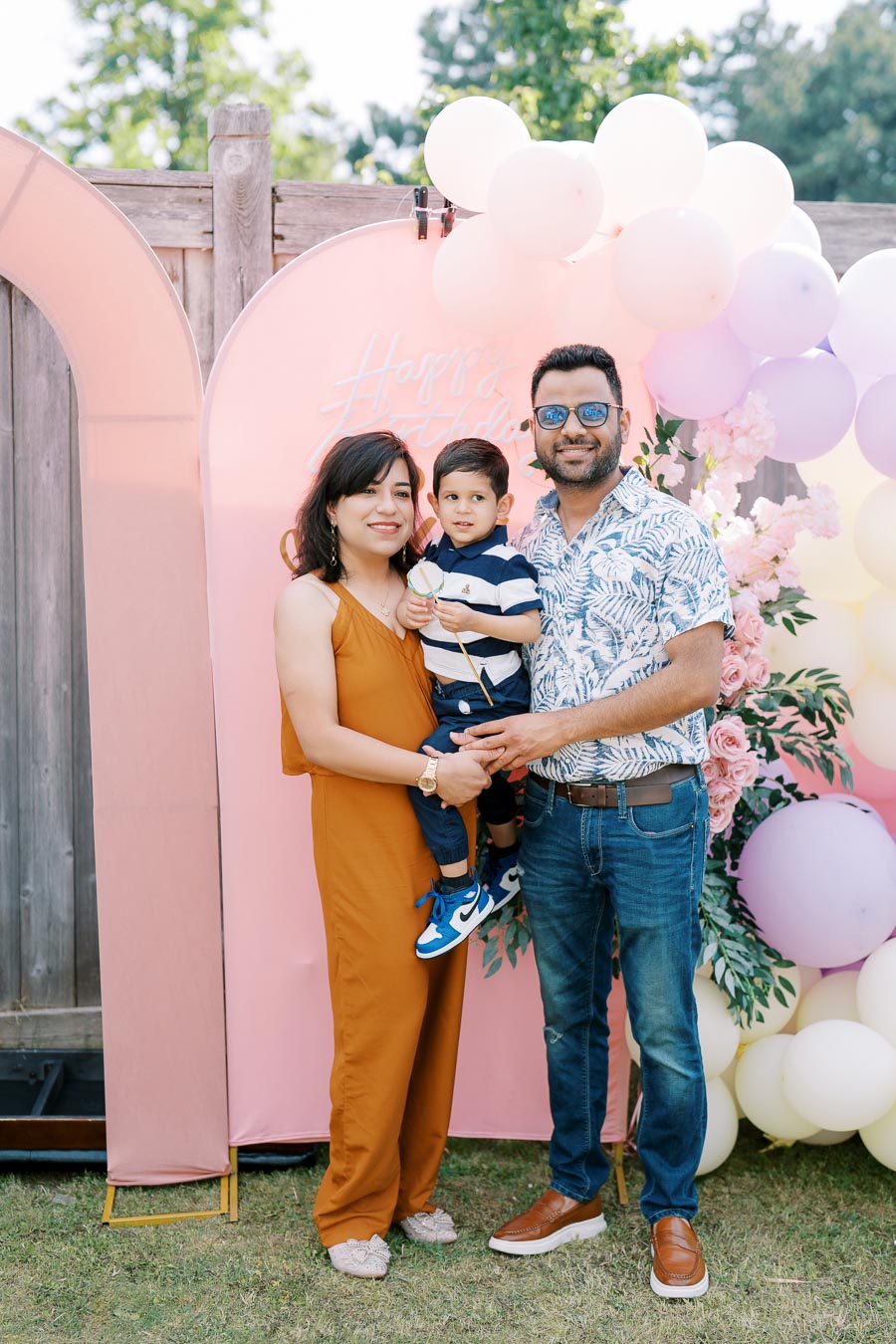 Family celebrating a birthday outdoors with colorful balloons and decorations, featuring a cheerful mother, father, and child posing in front of a pink backdrop that says Happy Birthday.