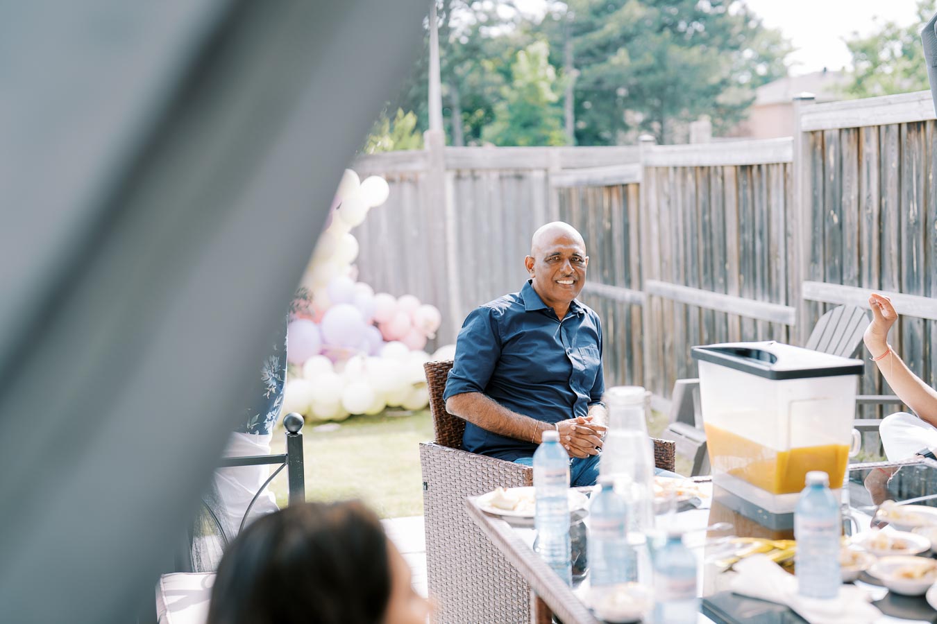 A man in a blue shirt smiling while seated at an outdoor gathering, surrounded by a table with water bottles and food, against a backdrop of pastel balloons and a wooden fence.
