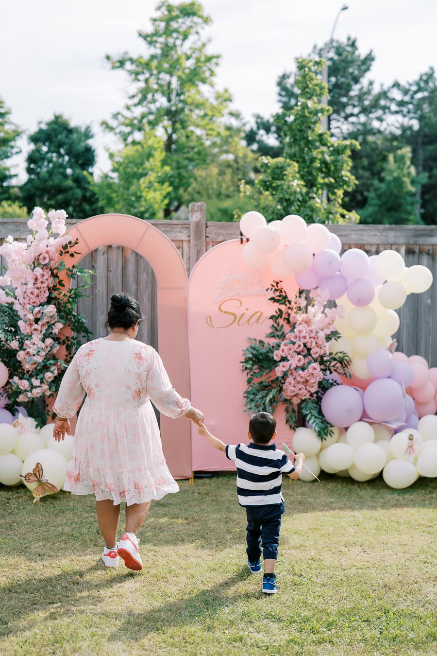 A woman in a floral dress holding hands with a young boy at an outdoor birthday party, walking towards a pastel pink and white balloon and flower arch backdrop.