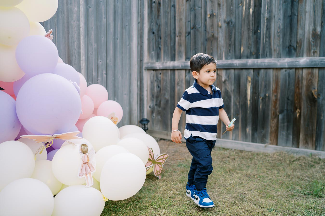 A young child in a striped shirt walks on grass beside a colorful balloon arrangement during an outdoor birthday party with a wooden fence in the background.