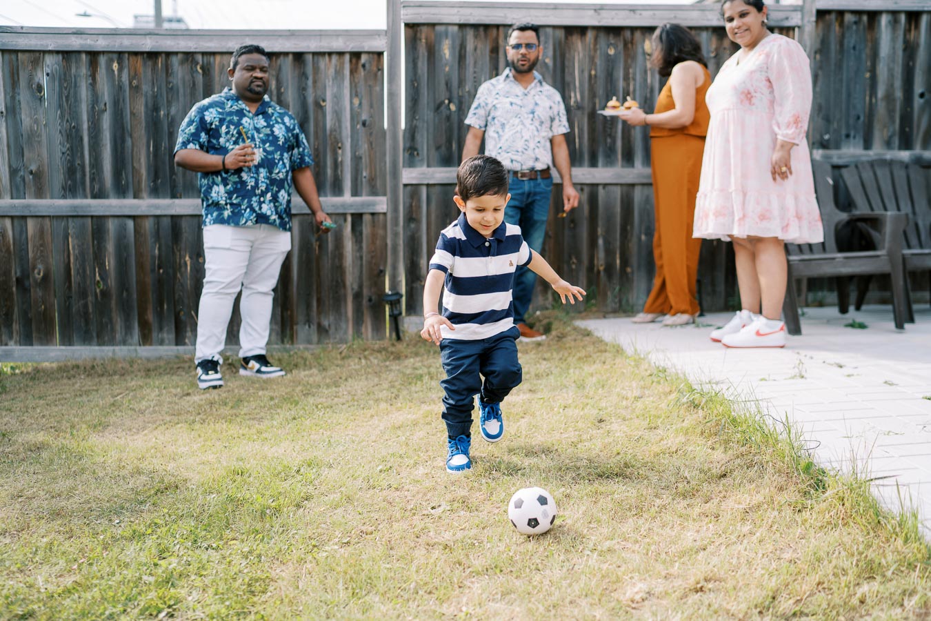 A young boy energetically plays soccer in a backyard, with adults socializing in the background during a sunny day gathering.