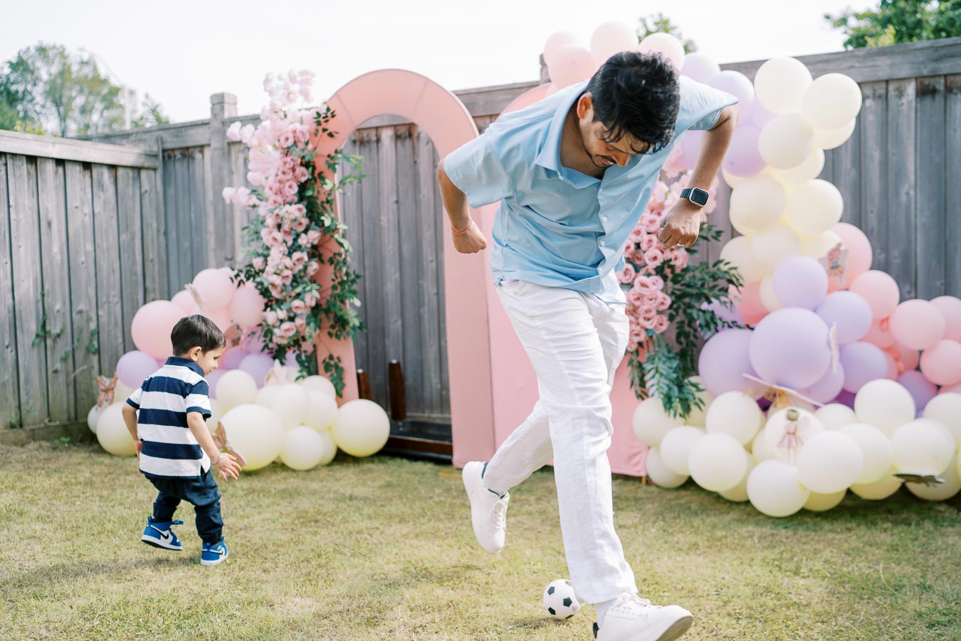 A man in a blue shirt and white pants kicking a small soccer ball in a garden decorated with pink and white balloons, alongside a child in a striped shirt.