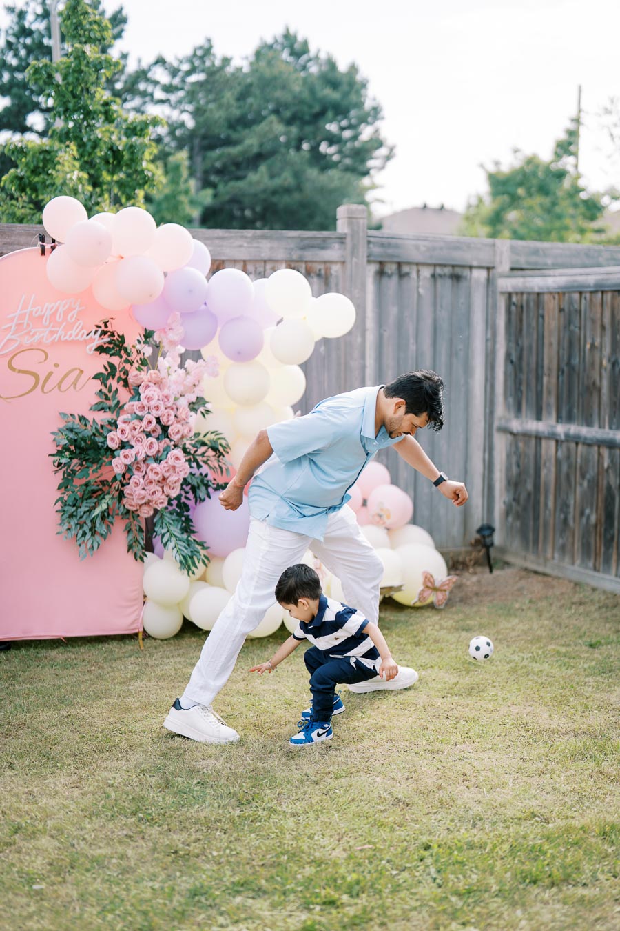 Father and young son playing soccer in a backyard decorated for a birthday party, with a pink backdrop, pastel balloons, and floral arrangements.