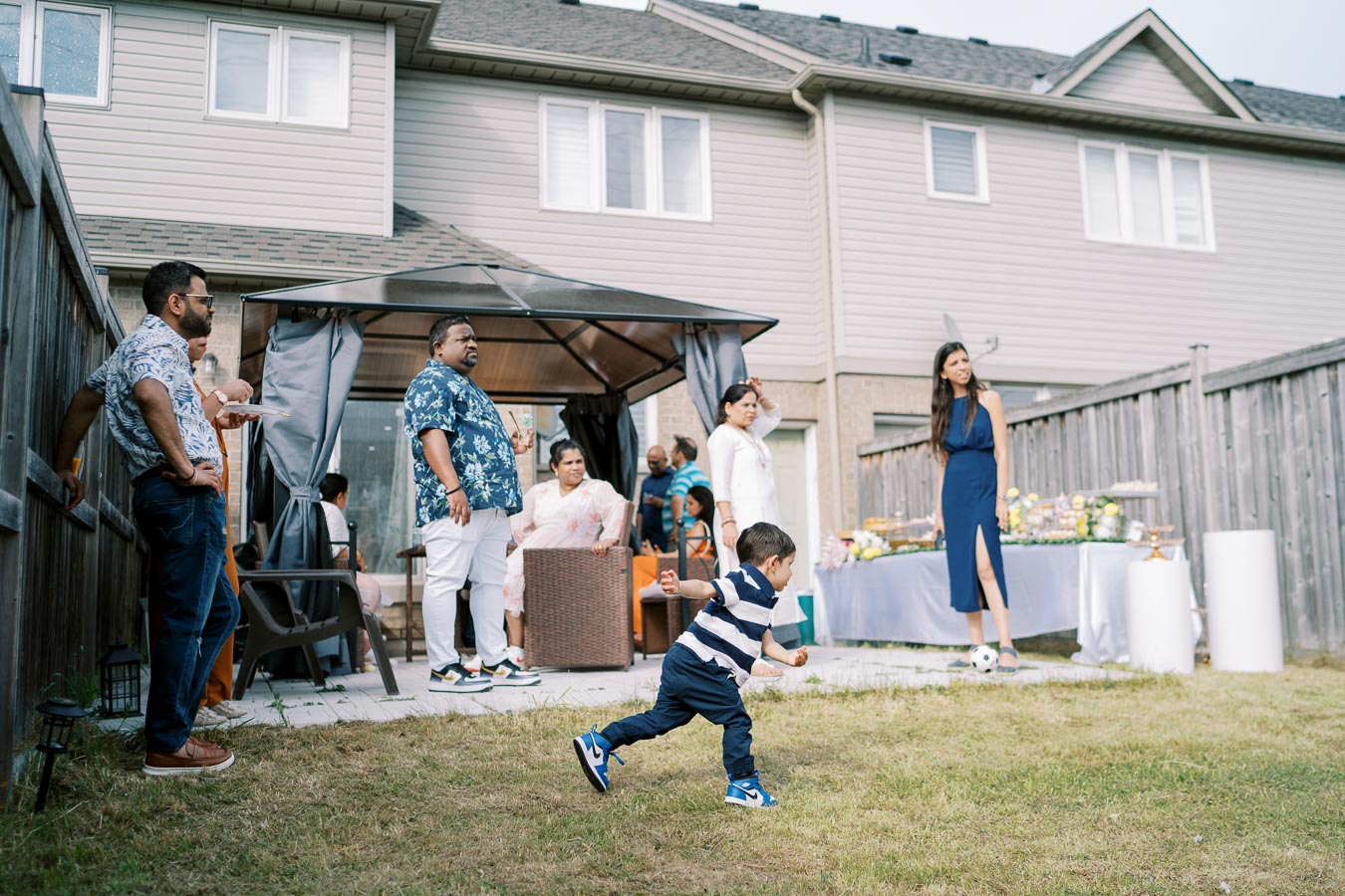 A lively backyard gathering with a young boy in a striped shirt running on the grass, surrounded by a group of adults chatting and relaxing under a canopy. A table in the background is set with food, indicating a festive occasion near a suburban house.