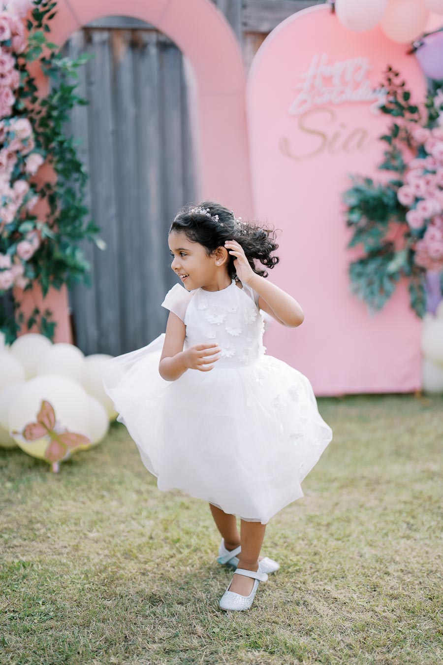 A joyful young girl in a white dress twirls at an outdoor birthday party decorated with pink floral arches and balloons.