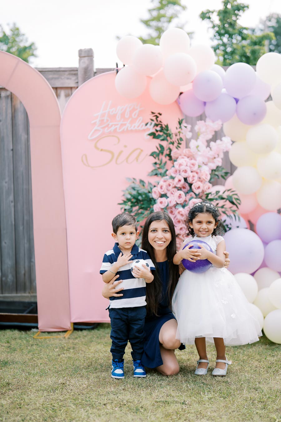 An adult and two children smiling at a birthday party, surrounded by pastel-colored balloons and flowers, with a pink birthday backdrop that says Happy Birthday.