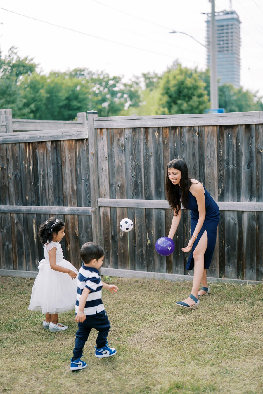 A woman in a navy dress plays with two young children, tossing colorful balls in a grassy backyard with a wooden fence in the background.