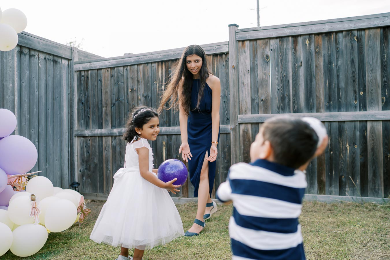 A woman in a blue dress playing a ball game with two children in a backyard decorated with purple and white balloons.