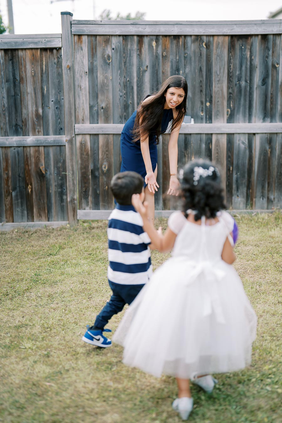 A woman in a blue dress smiling and playing with two children, a boy in a striped shirt and a girl in a white dress, in a grassy backyard with a wooden fence in the background.