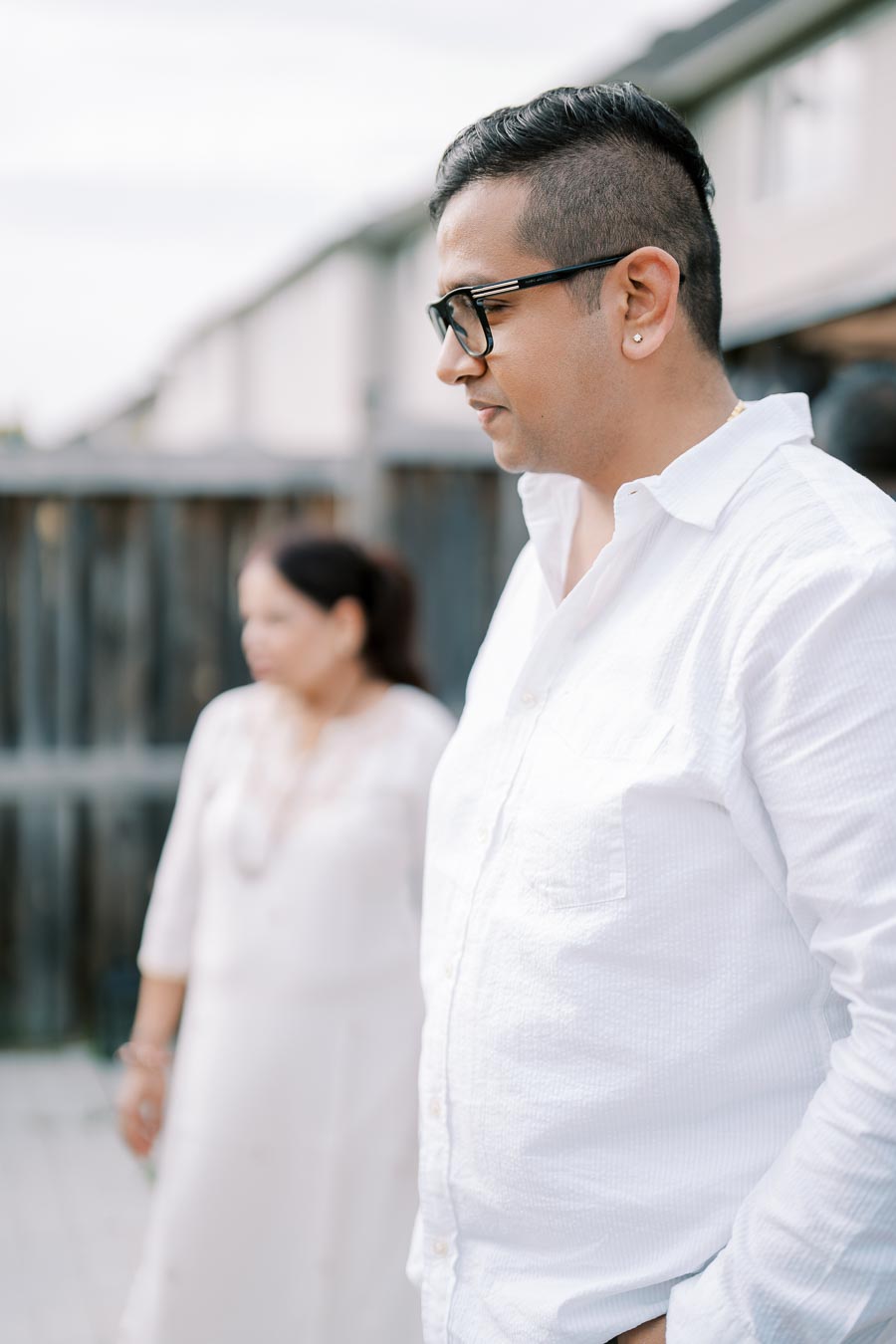 A person wearing glasses and a white shirt stands outdoors, looking to the side, with a blurred figure in the background, captured in a residential setting.
