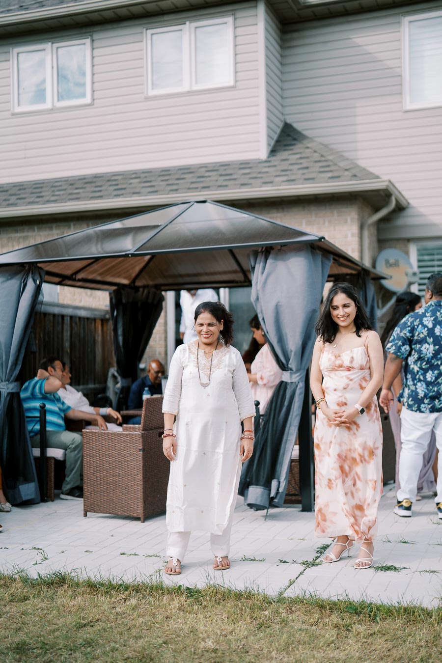 Two women standing in a backyard garden party, with a gazebo and guests in the background. One woman wears a white traditional outfit while the other wears a floral dress, creating a festive and celebratory atmosphere.