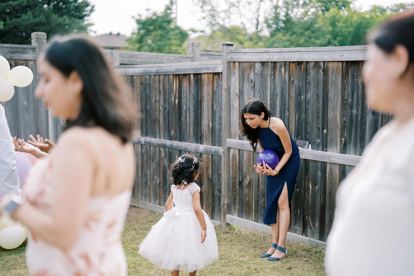 A woman in a navy blue dress is crouching to interact with a young girl in a white dress, holding a purple ball at an outdoor gathering. A wooden fence and green trees serve as the background, with blurred people visible in the foreground.