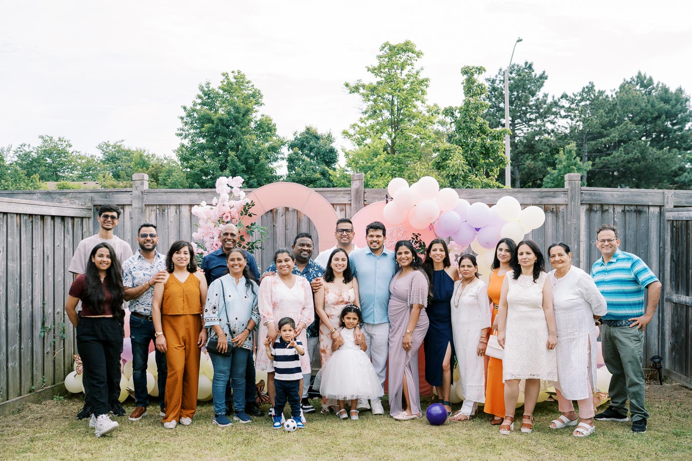 A large group of family and friends pose happily together in a backyard setting for a celebration, surrounded by colorful balloons and floral decorations.