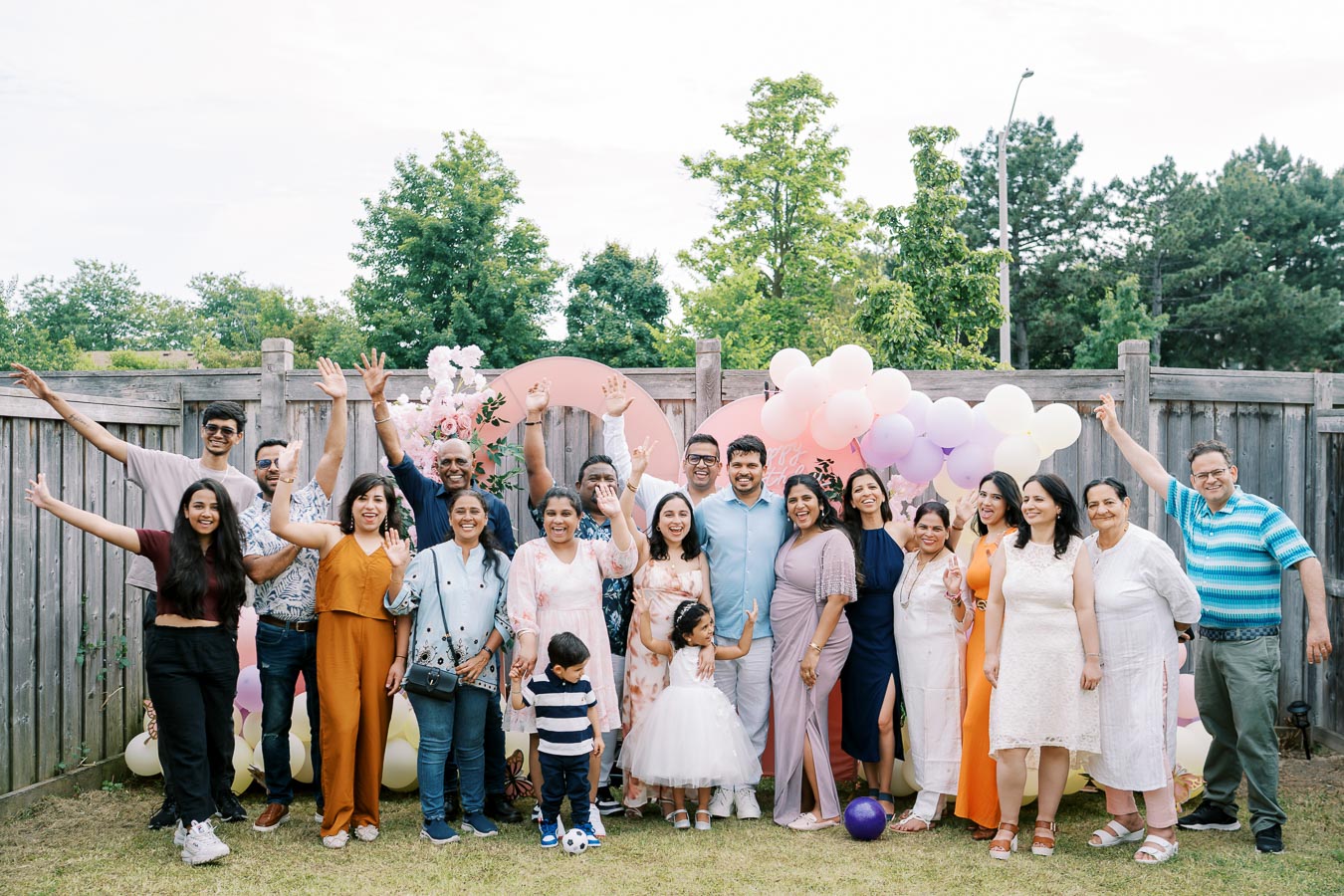 A diverse group of people celebrating a special occasion outdoors, posing in front of a decorated backdrop featuring pastel balloons and floral arrangements, with a wooden fence and lush trees in the background.