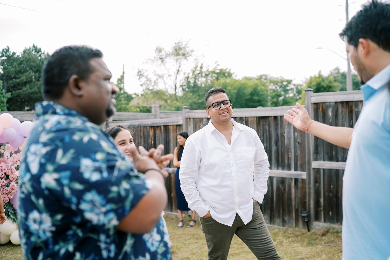 A group of people engaging in a friendly outdoor gathering in a backyard, with balloons and flowers in the background.