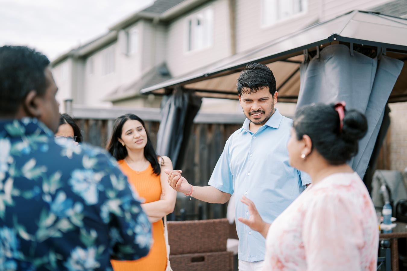 A group of people engaging in a lively conversation outdoors near a pergola on a sunny day, with houses visible in the background.