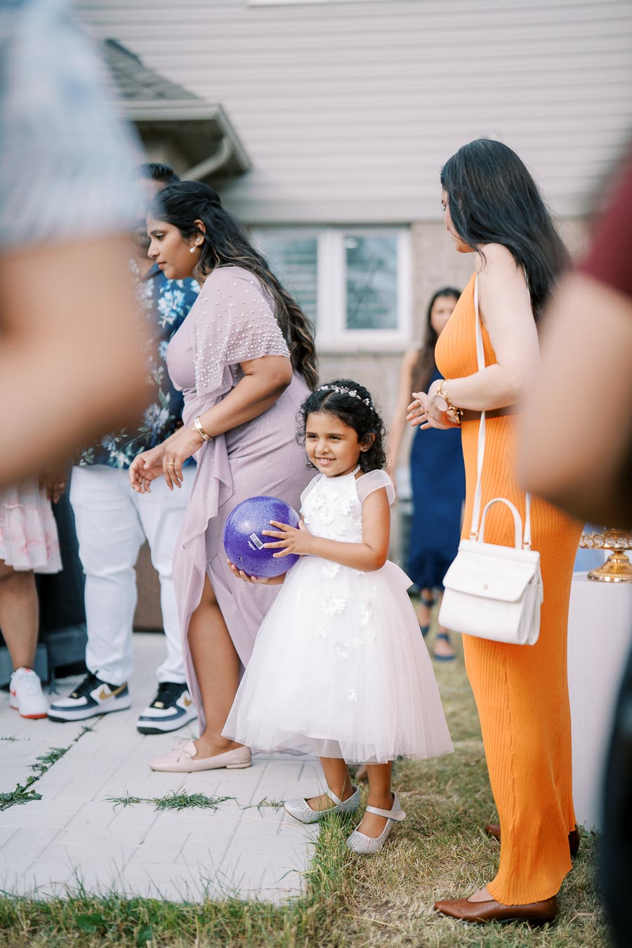 A young girl in a white dress holding a purple ball, surrounded by family and friends at an outdoor gathering. She is smiling and wearing a floral headband, while adults around her are engaged in conversation. The setting appears to be in a backyard with grass and a patio area.